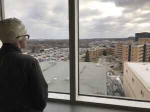 A teen in head bandages and glasses looks out a window overlooking hospital grounds.