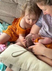 A young sibling looks on as a new mother breastfeeds her infant on a pillow.