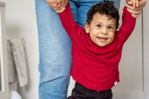 A curly haired child smiles while his parent holds his arms above his head, helping him walk.