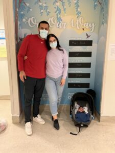 Parents pose in front of a NICU graduation sign with their infant son in a carrier on the ground next to them.