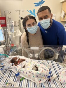 Two parents in face masks lean over their newborn's NICU bed.