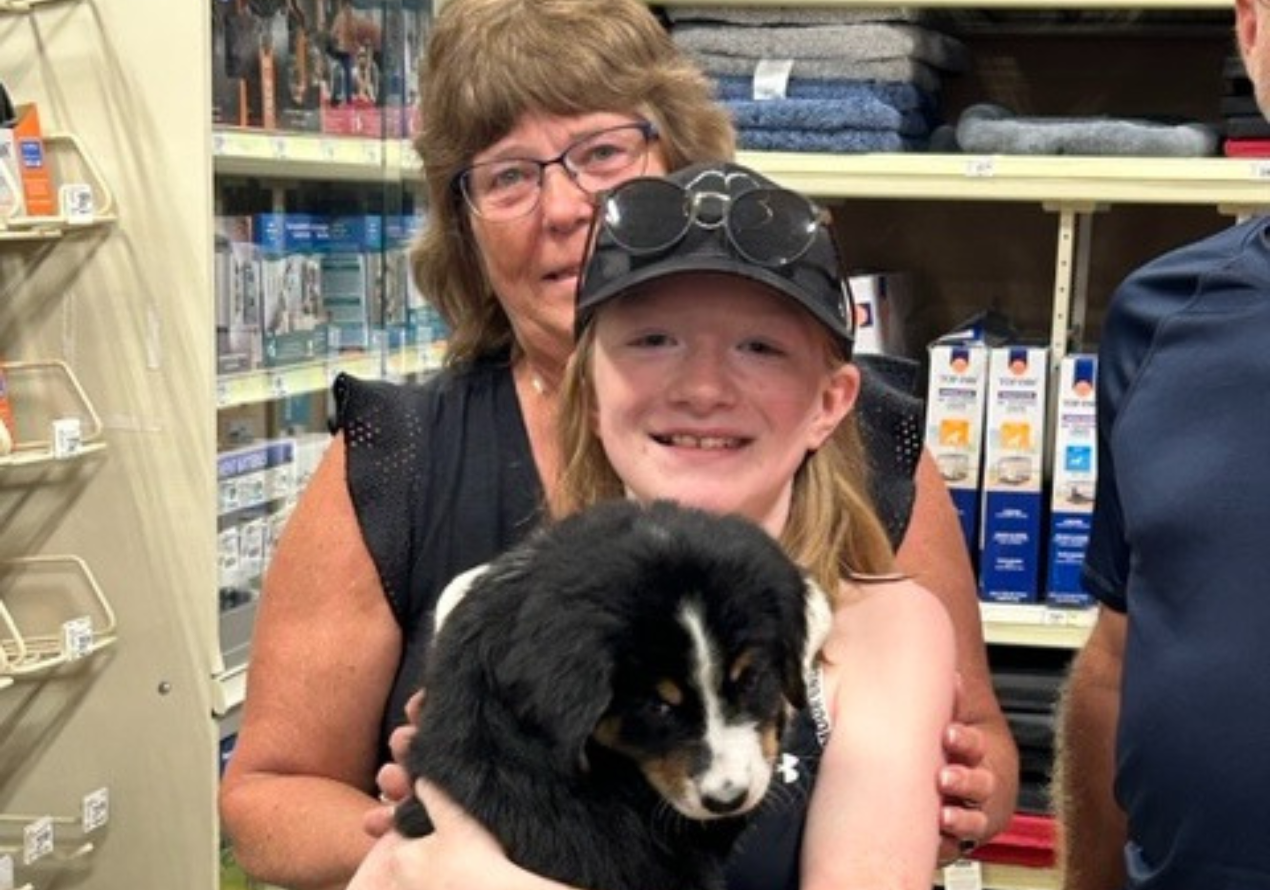 A woman stand behind a girl in a hat who is holding a black and white puppy in her arms.