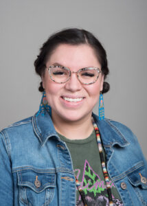 A headshot of Nicole Yawney, in which she wears a jean jacket glasses, and traditional earrings.