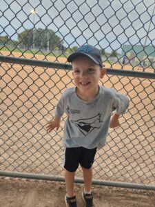 A boy in a gray Toronto Blue Jays shirt and a baseball cap leans back against a baseball backstop.