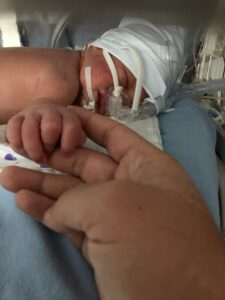 A hand reaches out to hold an infant in a NICU bed.