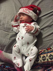 An infant in Christmas-themed onesie and hat sleeps peacefully.