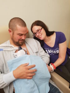 A father and mother pose while the father holds their infant, wrapped in a blue blanket, to his chest.