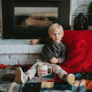 A boy with blond hair sits in front of a fireplace with a Christmas-themed mug between his legs.